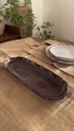 Homura Artisan Platter Captivating with its rich burgundy glaze and gently scalloped rim, this expansive stoneware platter brings a touch of dramatic warmth to your table. Its low, sleek profile makes it an exquisite backdrop for artistic food presentation or a striking accent piece for grand hosting.