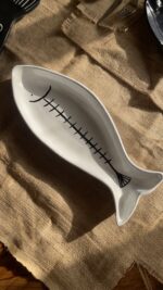 Azura Serving Platter Crafted from durable stoneware, this beautiful fish-shaped serving platter stands out with its clean white glaze and artistic hand-painted black fishbone design. It is perfect for adding a stylish sea-inspired vibe to any meal.